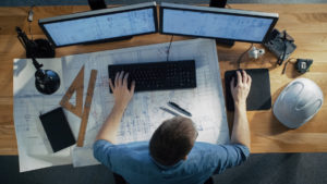 Draufsicht des Architekturingenieurs Working on His Blueprints, Tablet-Computer halten und auch Tischrechner verwenden. Sein Schreibtisch ist voller nützlicher Gegenstände und Abendsonne.