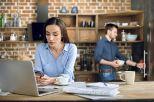 Frau, die Freelancer ist, sitzt mit Kaffee, Laptop un Smartphone am Küchentisch, im Hintergrund befindet sich ein Mann