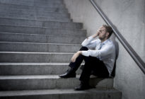 Mann im Business-Look sitzt auf Treppe und hält verzweifelt rechte Hand in seinem Gesicht.