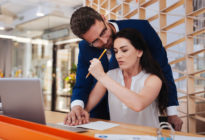 Mann legt am Arbeitsplatz seine Hand auf die einer Frau, die am Schreibtisch mit Laptop sitzt. Sie wehrt sich mit einer Faust, in der sie einen Bleistift hält.