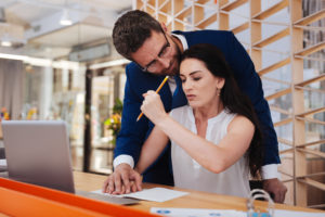 Mann legt am Arbeitsplatz seine Hand auf die einer Frau, die am Schreibtisch mit Laptop sitzt. Sie wehrt sich mit einer Faust, in der sie einen Bleistift hält.