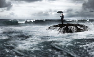 Krise: Business-Mann mit Regenschirm steht auf kleiner Felsen-Insel im reißenden Meer