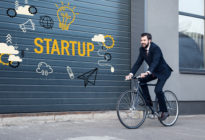 Fahrradfahrer im Business-Look, der an Grarage mit Aufschrift Startup vorbeiradelt