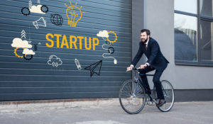 Fahrradfahrer im Business-Look, der an Grarage mit Aufschrift Startup vorbeiradelt