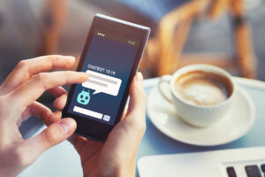 Hand mit Smartphone und Chatbot-Anzeige, sowie Kaffeetasse daneben