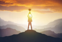 Silhouette eines Mannes auf einem Berg im Sonnenuntergang, der einen Pokal hochhält